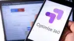 Usuario sosteniendo un teléfono móvil con pantalla que muestra el logo de Google Optimize 360. En segundo plano, una laptop muestra estadísticas difusas de un sitio web en Google.