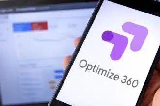 Usuario sosteniendo un teléfono móvil con pantalla que muestra el logo de Google Optimize 360. En segundo plano, una laptop muestra estadísticas difusas de un sitio web en Google.