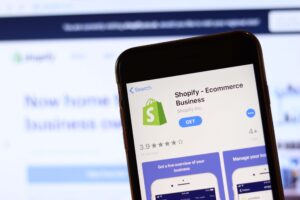 Teléfono con icono de Shopify en pantalla de cerca con sitio web borroso en portátil.